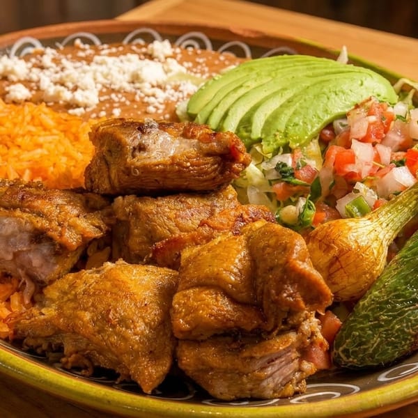 A colorful Mexican dish with carnitas, rice, refried beans, avocado, pico de gallo, and grilled vegetables.