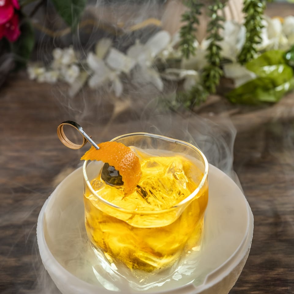 A smoking cocktail with orange peel and cherry garnish sits on a wooden table with flowers.