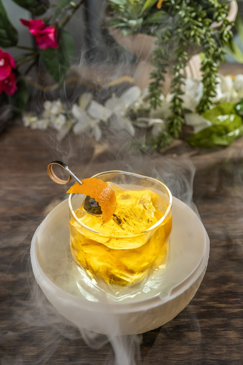 A smoking cocktail with orange peel and cherry garnish sits on a wooden table with flowers.