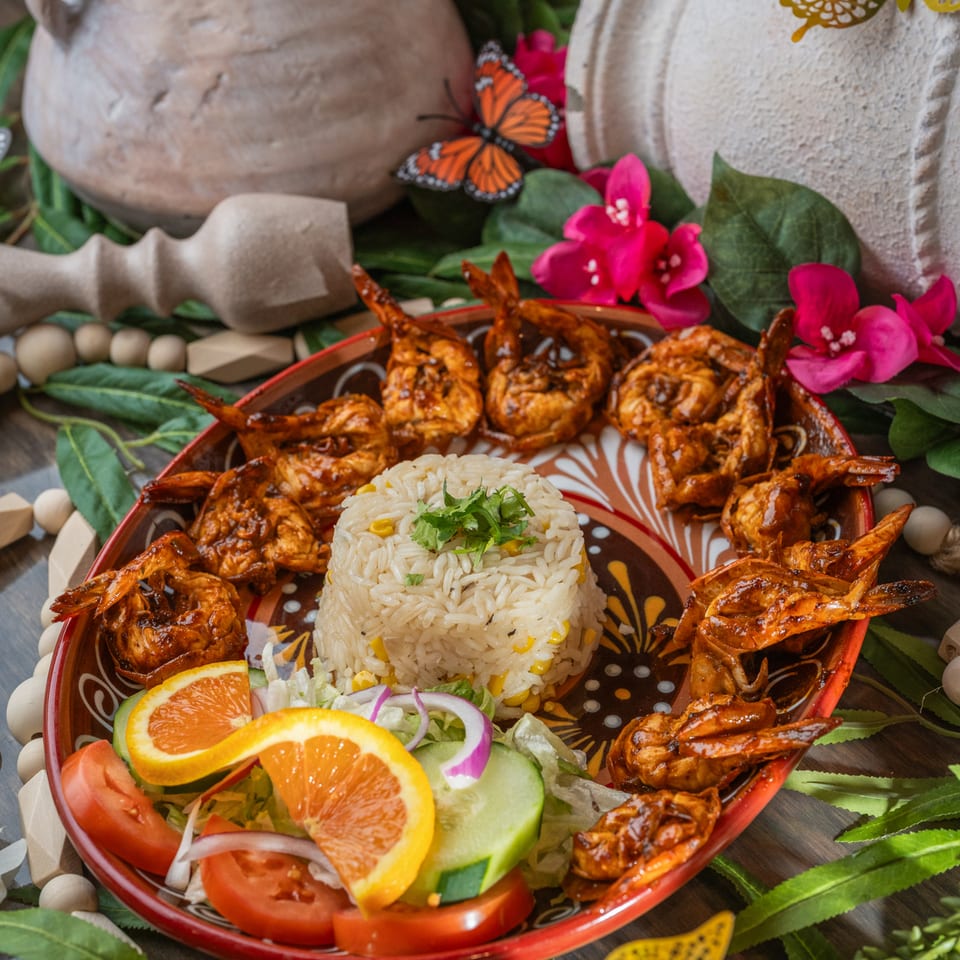 Platter of glazed grilled shrimp surrounding rice, salad, and orange slices, styled with flowers, greenery, pottery, and decorative butterflies.