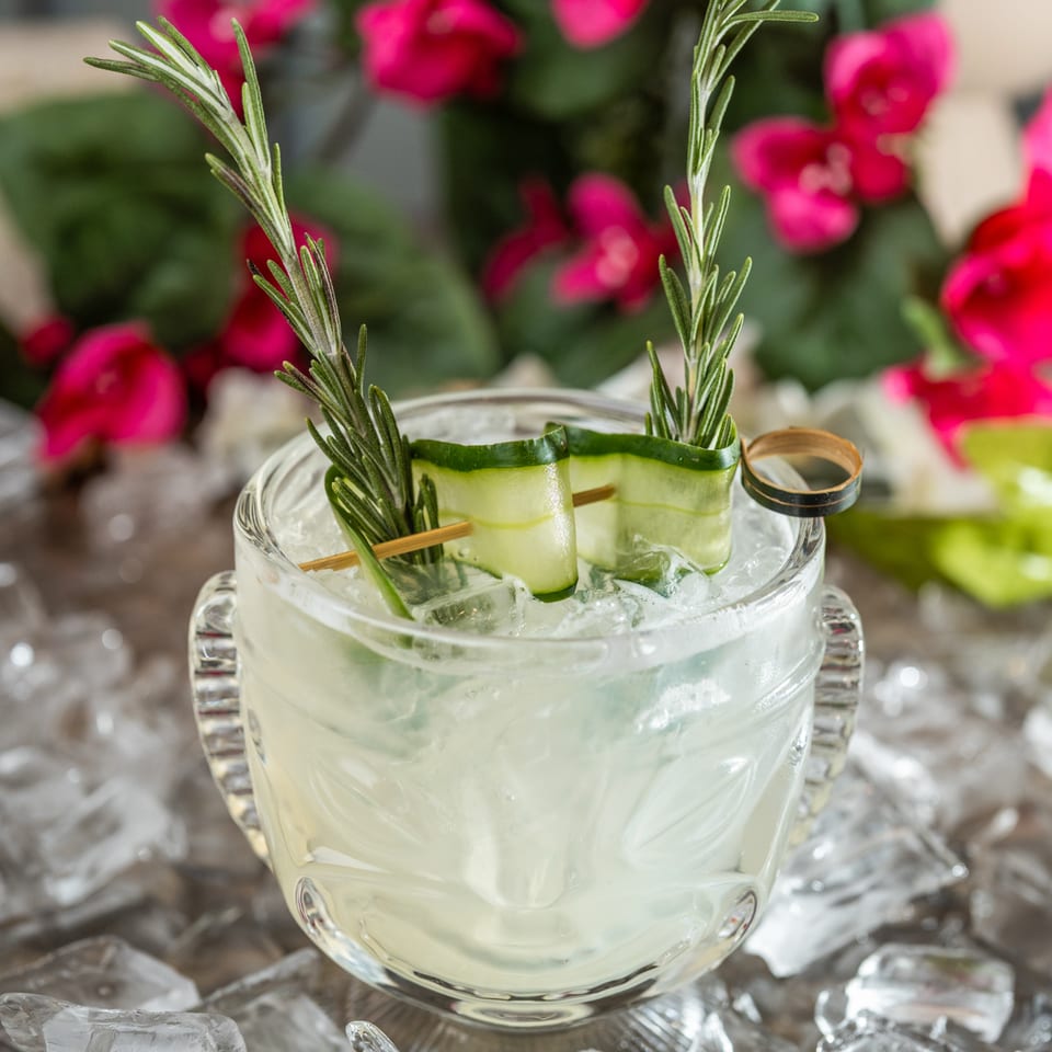 Icy pale cocktail in a glass mug garnished with cucumber ribbons and rosemary, surrounded by ice and pink flowers.