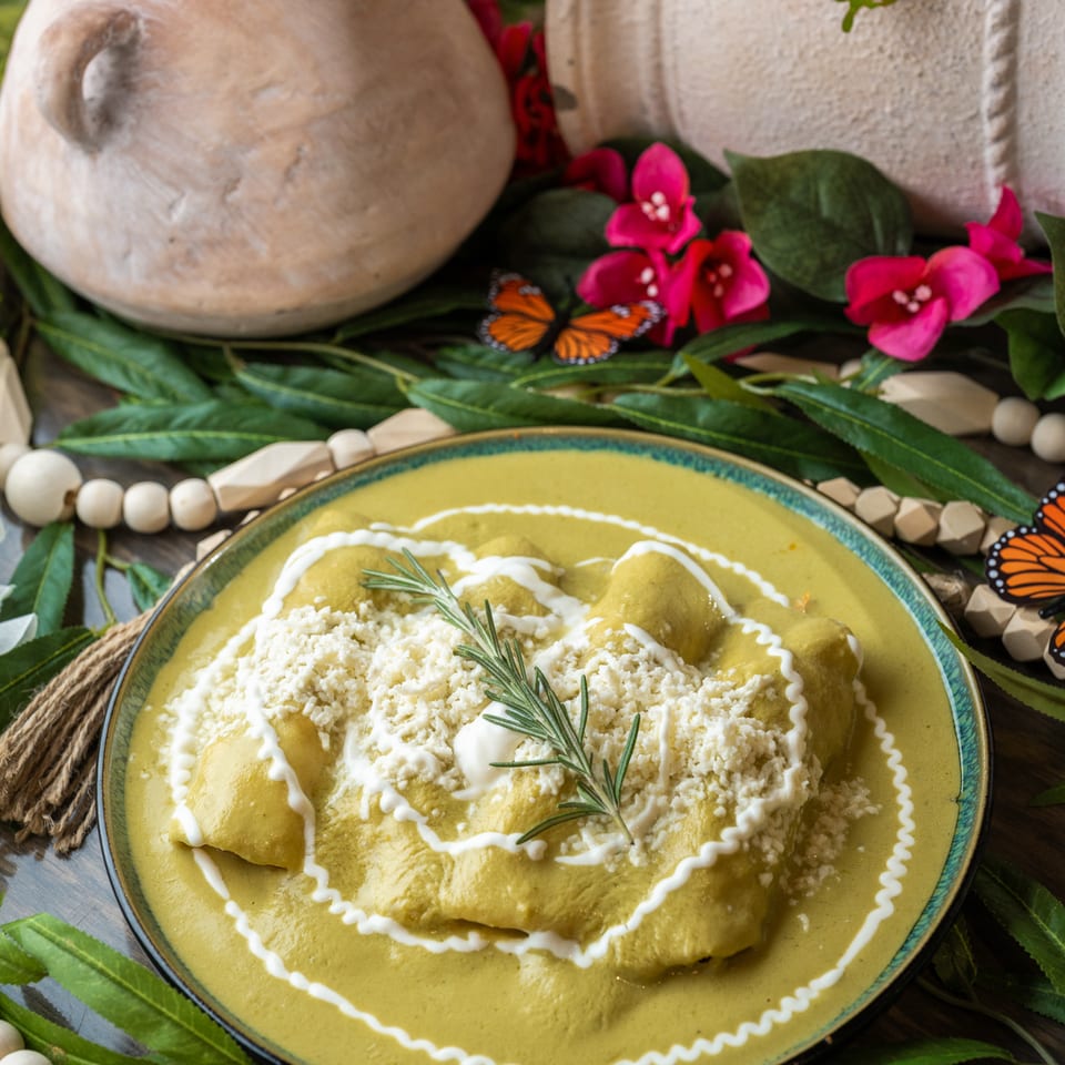 Plate of enchiladas covered in green sauce, crema, crumbled cheese, and rosemary, surrounded by flowers, leaves, pottery, and butterfly decorations.