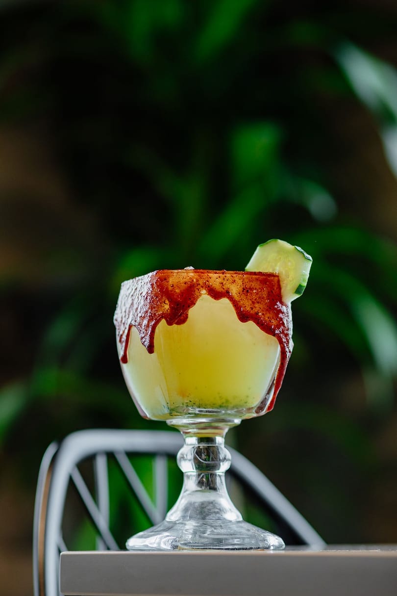 A yellow cocktail in a goblet, rimmed with red chili and garnished with a cucumber slice.
