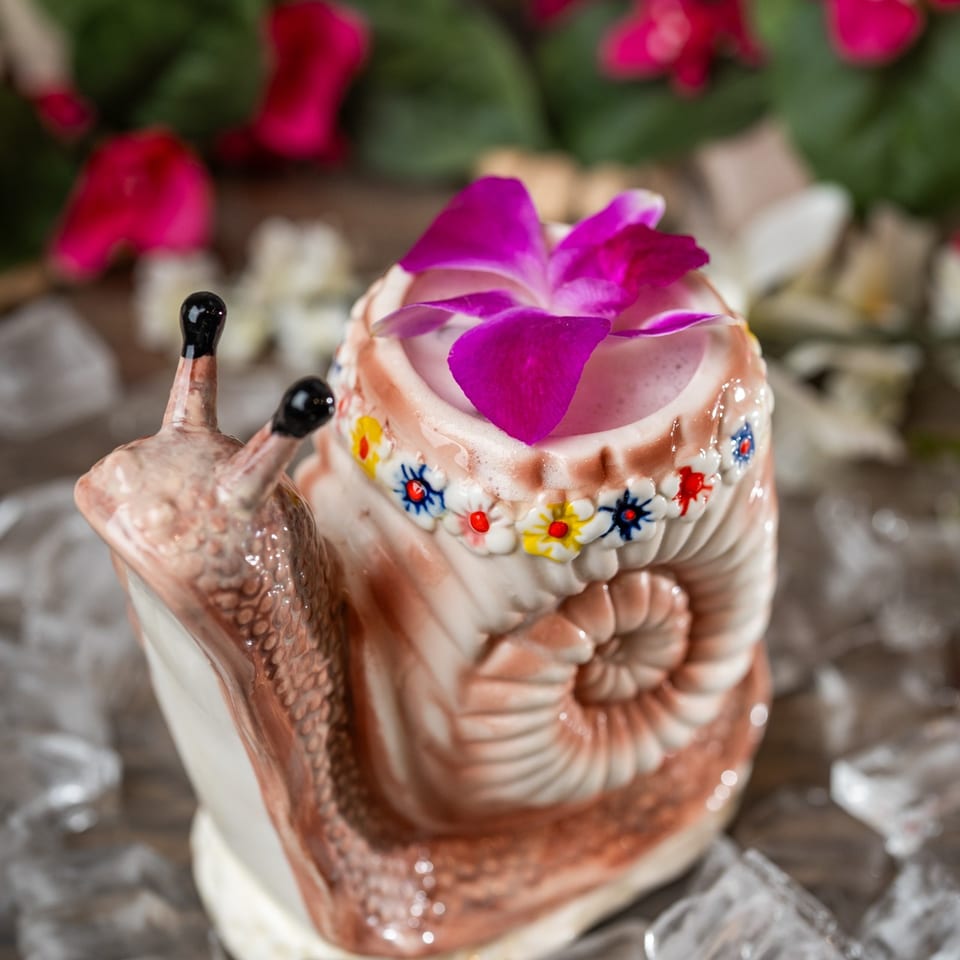 A snail-shaped cocktail mug with a pink drink and purple orchid garnish, surrounded by ice.