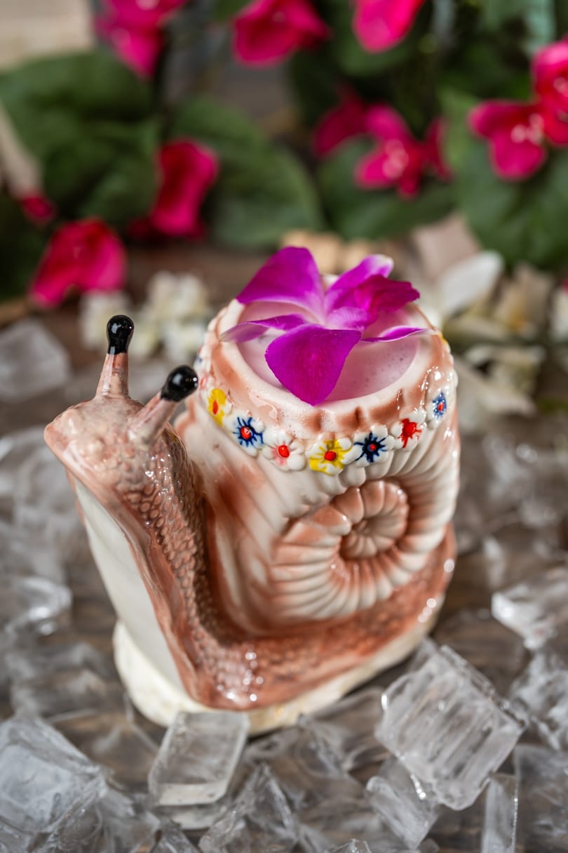 A snail-shaped cocktail mug with a pink drink and purple orchid garnish, surrounded by ice.