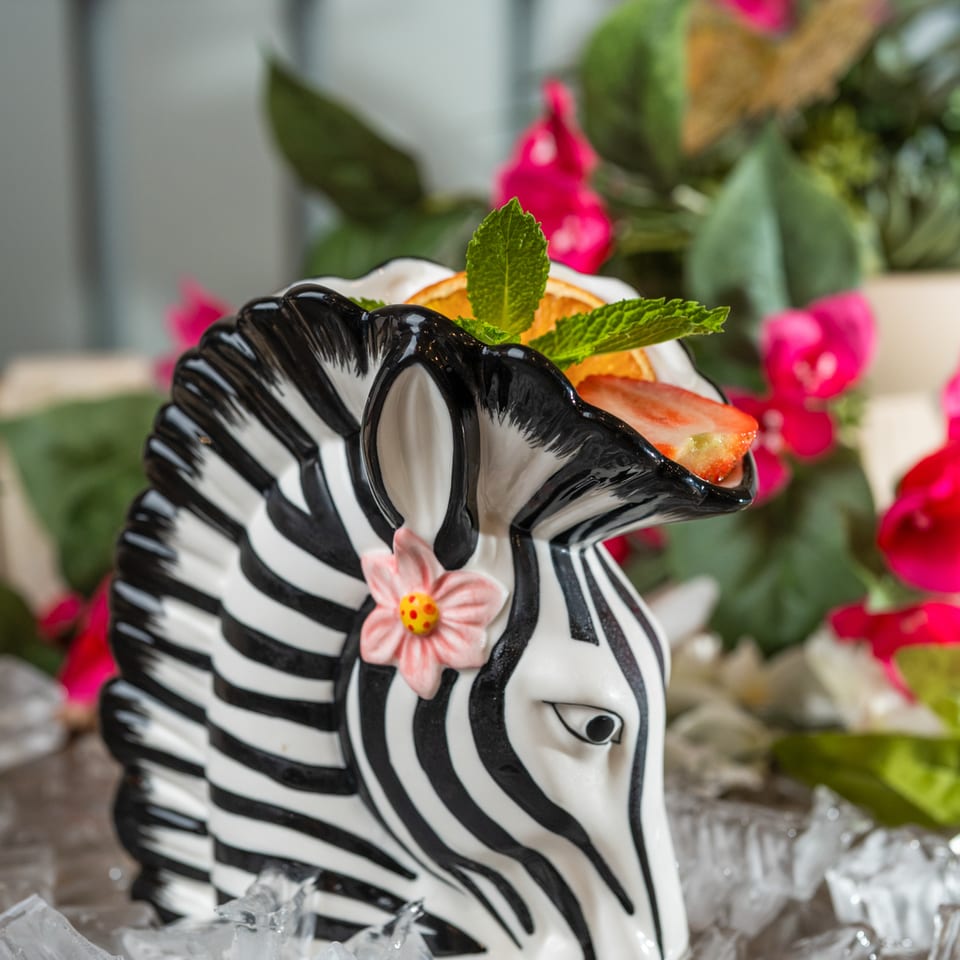 Zebra-shaped ceramic bowl holds fruit and mint on ice, surrounded by colorful flowers.