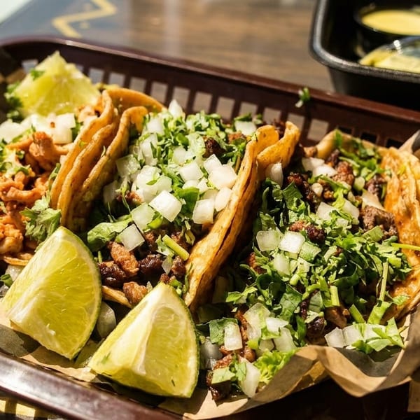 Three street tacos with meat, onions, cilantro, and lime wedges in a basket with sauces.