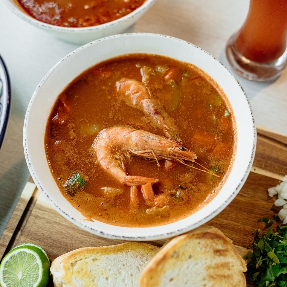 Bowl of shrimp soup with vegetables, bread, lime, radishes, cilantro, and chili flakes on a wooden table.