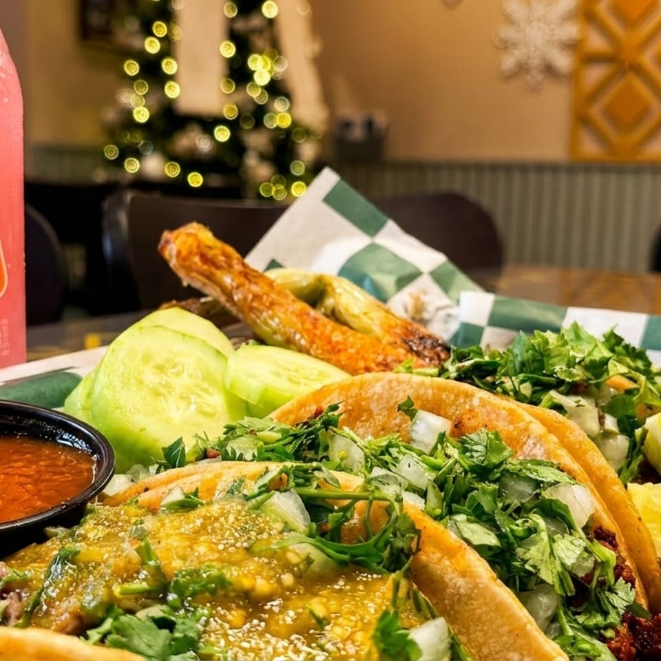 A tray of tacos with a side of salsa, limes, cucumbers, and a pink Jarritos soda.