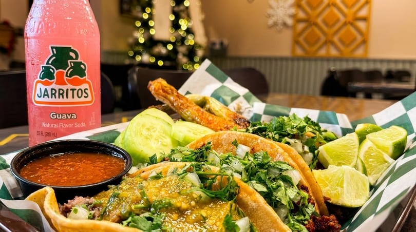 A tray of tacos with a side of salsa, limes, cucumbers, and a pink Jarritos soda.