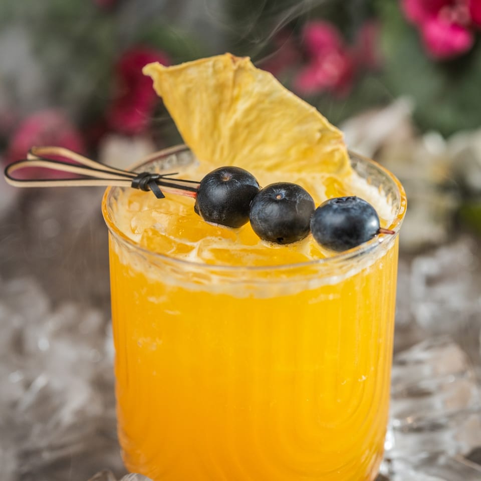 A vibrant orange cocktail with blueberries and a pineapple slice, surrounded by ice and mist.
