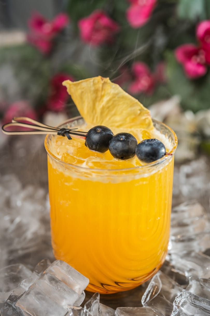 A vibrant orange cocktail with blueberries and a pineapple slice, surrounded by ice and mist.