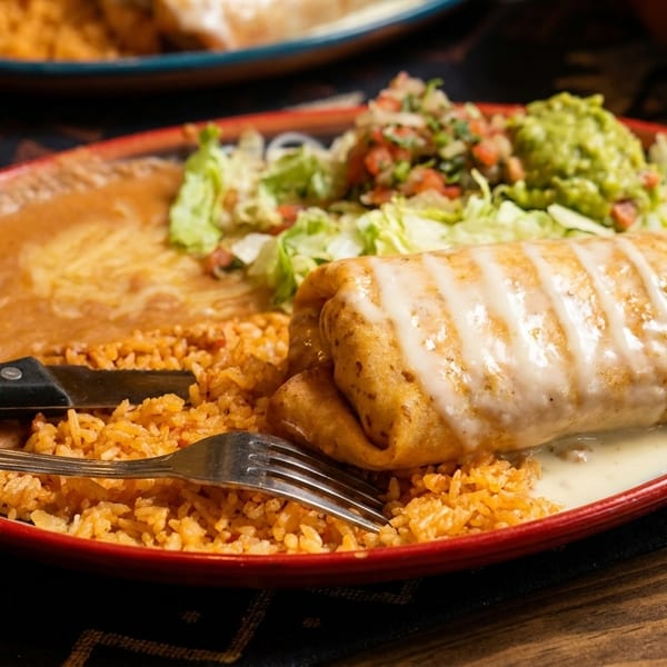 A large burrito covered in white sauce, with rice, refried beans, lettuce, pico de gallo, and guacamole.